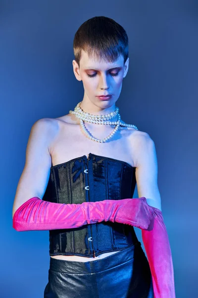 queer person in black corset and pearl necklace wearing pink gloves on blue backdrop, edgy style