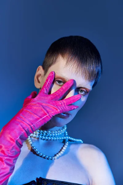 queer model in pearl necklace covering face with hand in pink glove on blue backdrop, edgy style