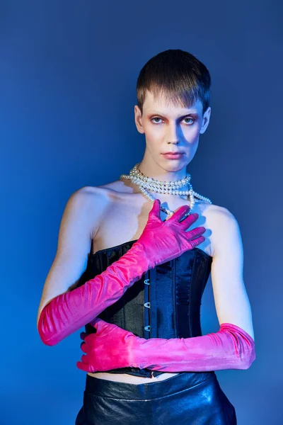 nonbinary person in black corset and pearl necklace posing in pink gloves on blue backdrop, queer