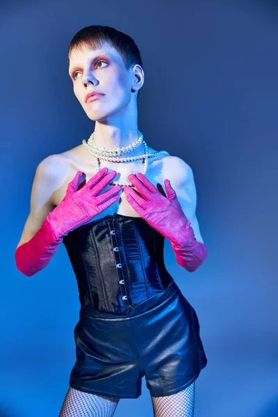 nonbinary person in corset and pearl necklace posing on blue backdrop, queer model looking away