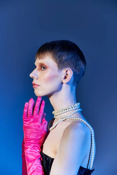 side view, nonbinary person in corset and pink gloves posing on blue backdrop, queer model