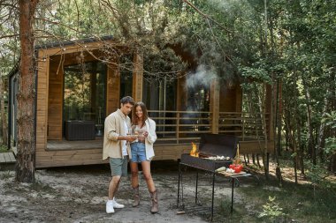Gülümseyen romantik çift şarapla kadeh kaldırıyor ve barbekü ve açık havada tatil evinin yanında duruyor.