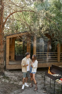 Şarabı olan romantik çift arka planda bbq ve yazlığın yanında dikilirken konuşuyor.