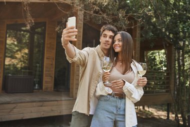 Şık bir adam, arka planda yazlık evin yanında kız arkadaşıyla birlikte akıllı telefondan selfie çekiyor.