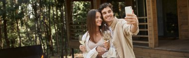 Şarabı olan gülümseyen çift, arka planda BBC ve yazlık evin yakınındaki akıllı telefondan selfie çekiyor.
