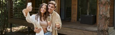 Şarapla gülümseyen romantik bir çift. Akıllı telefondan selfie çekiyor. Bulanık yazlık evin yanında, pankartta.