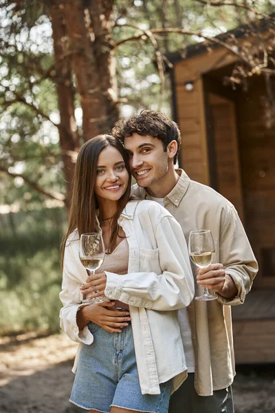 Smiling couple with wine hugging and looking at camera together near blurred vacation house