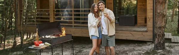 一对浪漫的夫妇在烧烤和度假屋附近举着酒 看着相机 高举横幅 — 图库照片