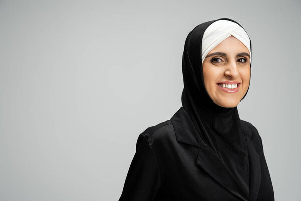 happy muslim businesswoman in hijab and black jacket smiling at camera on grey, headshot