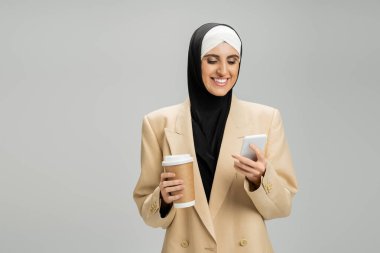 Kafiyeli ve ceketli, akıllı telefon ve kahveyle gri üzerine gitmek için motive olmuş Müslüman iş kadını.