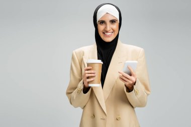 Bej ceketli ve tesettürlü mutlu Müslüman iş kadını elinde kahve ve gri üzerinde akıllı telefon tutuyor.