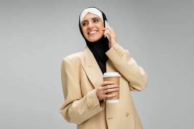 Neşeli, modaya uyan Müslüman iş kadını elinde kahve, akıllı telefonuyla konuşuyor, gözlerini griden ayırmıyor.