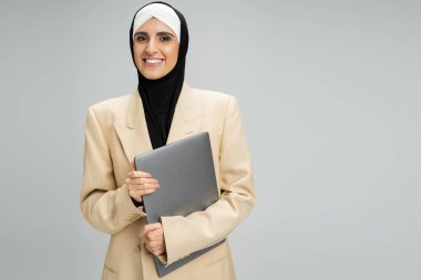 Spor ceketli, tesettürlü, tesettürlü Müslüman bir iş kadınının gri dizüstü bilgisayarı tutuşu, başarı.