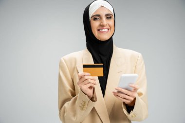 Cep telefonu ve kredi kartıyla gri kameraya gülümseyen şık bir Müslüman iş kadını.