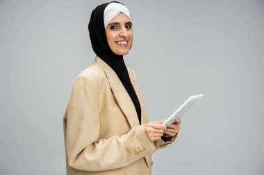 Modern Müslüman kadın tesettüre ve şık iş kıyafetlerine sahip dijital tablet tutuyor ve gri üzerine gülümsüyor.