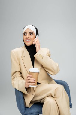 Gülümseyen, şık Müslüman iş kadını elinde kağıt, gri koltukta akıllı telefonla konuşuyor.
