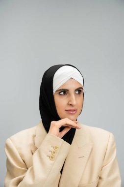 Başörtüsü ve ceketli zarif Müslüman iş kadını, çenesine yakın eli griye bakıyor.