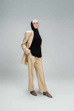 confident muslin woman in hijab and stylish business attire looking at camera on grey, full length clipart