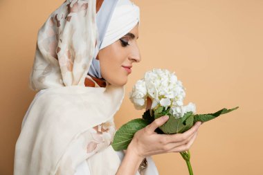 Beyaz ortanca çiçeğini bej, kapalı gözlerle tutan şık başörtüsü takan gülümseyen Müslüman kadın.