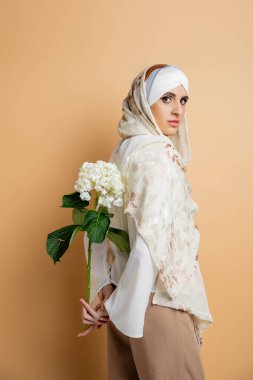 Beyaz ortanca çiçeğiyle şık giyinmiş çekici Müslüman kadın bej renkli kameraya bakıyor.