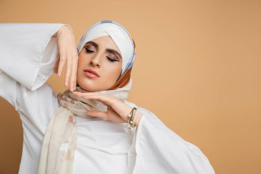 portrait of sensual muslim woman in silk scarf posing with closed eyes and hands near face on beige clipart