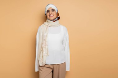 Beyaz bluzlu, ipek eşarplı, bej renkli kameraya gülümseyen neşeli Müslüman kadın.