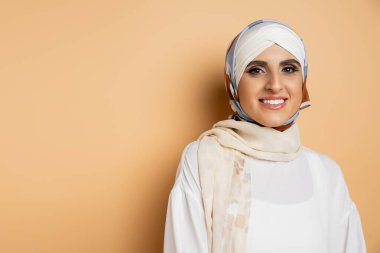 happy muslim woman with makeup wearing silk scarf and white blouse, smiling at camera on beige clipart