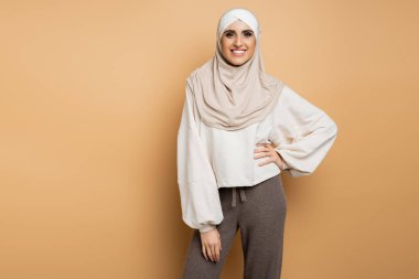 tesettürlü neşeli müslüman kadın ve modaya uygun günlük giysiler bej rengi elbiseler içinde elleri kalçasında duruyor.