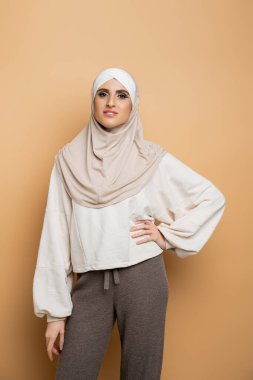 Mutlu Müslüman kadın tesettürlü ve şık gündelik giysiler içinde elleri bej üzerinde poz veriyor.