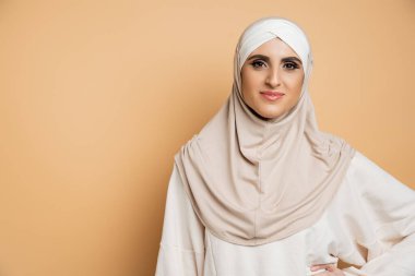 tesettürlü, eşofmanlı, eli belinde poz veren bej renkli kameraya bakan pozitif müslüman kadın.
