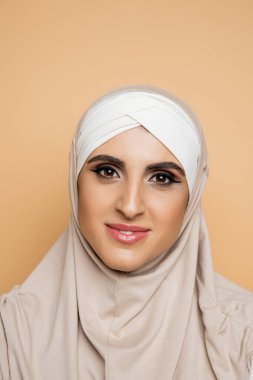 Makyajlı, tesettürlü ve bej renkli kameraya bakan neşeli Müslüman kadın portresi