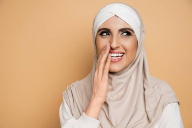 Heyecanlı, stil sahibi, tesettürlü Müslüman bir kadın. Ağzını eliyle kapatıyor, başka yere bakıyor ve bej rengi elbiseyle gülüyor.