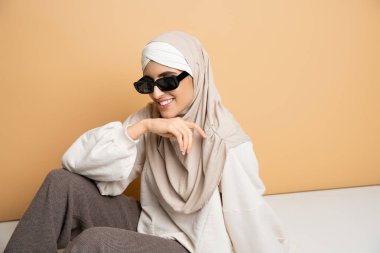 Mutlu Müslüman kadın tesettürlü, güneş gözlüklü ve modaya uygun giyinmiş bej rengi üzerinde oturuyor ve gülümsüyor.