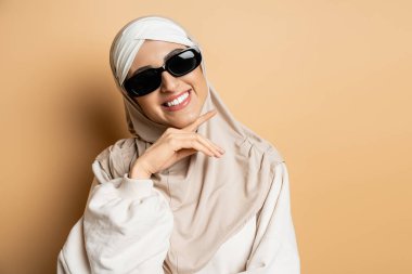 joyful muslim woman in hijab and trendy sunglasses posing with hand near chin and smiling on beige clipart