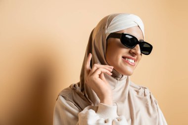 modern and smiling muslim woman in hijab and trendy sunglasses looking away while posing on beige clipart