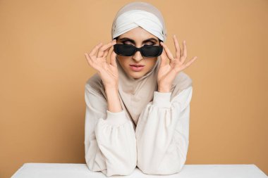 Başörtüsü ve sweatshirt giyen modaya uyan Müslüman kadın beyaz masada oturuyor, bej rengi güneş gözlüğünü çıkarıyor.