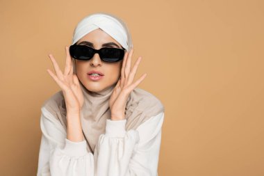 modern muslim woman in hijab and white sweatshirt touching stylish sunglasses while posing on beige clipart