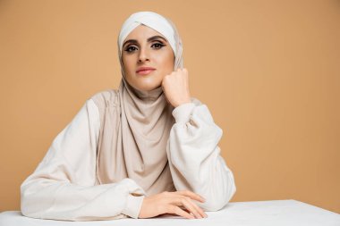 tesettürlü ve kazaklı bir müslüman kadın masada oturuyor, bej rengi giyinmiş kameraman kadına bakıyor.