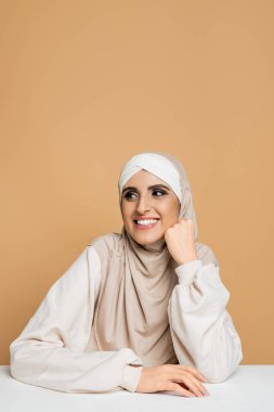 Beyaz tişörtlü, tesettürlü, kaygısız Müslüman kadın beyaz masada oturuyor ve bej rengine bakıyor.