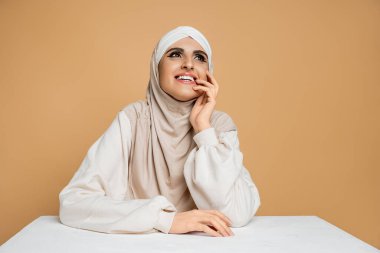 Modern Müslüman kadın tesettürlü ve sweatshirt 'lü beyaz masada oturuyor ve bej renge bakıyor.