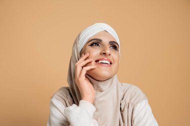 Beyaz kazaklı, tesettürlü ve tesettürlü heyecanlı müslüman kadın yüzünün yanında el ele tutuşup bej renge bakıyor.