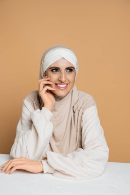 Beyaz kazaklı ve tesettürlü neşeli Müslüman kadın bej rengi masada otururken başını çeviriyor.