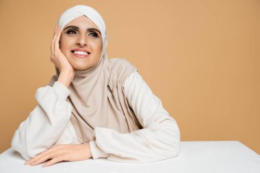 Şık eşofman altı ve tesettürlü mutlu ve rüya gibi Müslüman kadın bej üzerine beyaz masaya bakıyor.