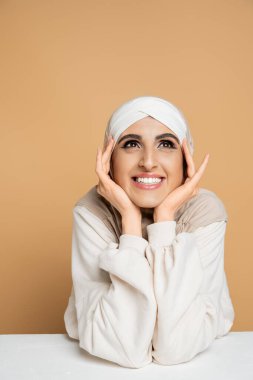 happy and dreamy muslim woman in hijab holding hands near face and looking away at table on beige clipart