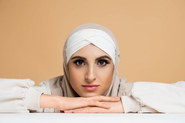 attractive muslim woman with makeup, sitting at white table in hijab and looking at camera on beige clipart