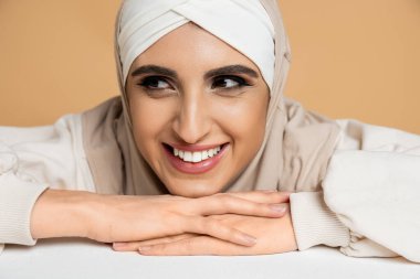 tesettürlü neşeli müslüman kadın portresi beyaz masada oturuyor ve bej renge bakıyor.
