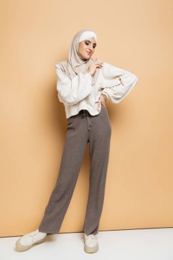 moda müslüman kadın tesettürlü, beyaz kazaklı ve gri pantolonlu, elleri bej rengi kalçasında poz veren