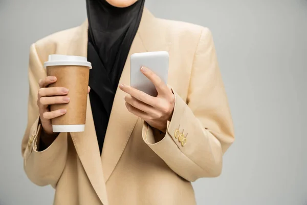 stock image cropped view of muslim businesswoman in beige blazer, with mobile phone and takeaway drink on grey