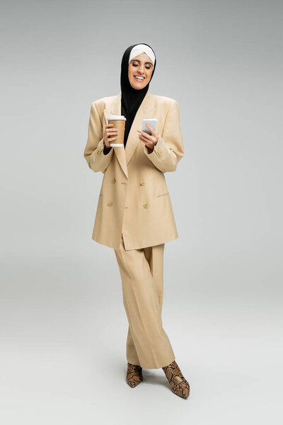 successful muslim businesswoman in hijab and beige suit holding paper cup and mobile phone on grey