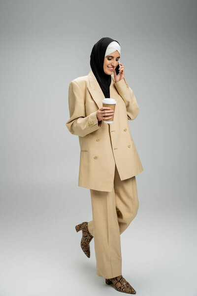muslim businesswoman in suit and hijab holding paper, talking on smartphone on grey, full length